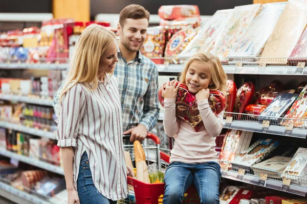 Sorridente bambina seduta su un carrello della spesa e scegliere caramelle con i suoi genitori al supermercato — Foto Stock