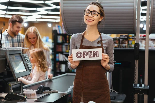 Portrait of friendly woman cashier holding open sign — Stock Photo, Image