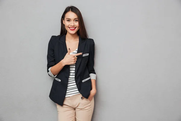 Sonriente asiático mujer de negocios con el brazo en el bolsillo apuntando lejos —  Fotos de Stock