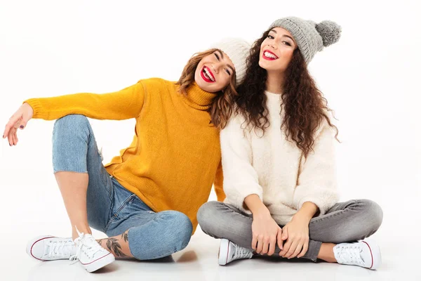 Две счастливые девушки в свитерах и шляпах сидят на полу — стоковое фото