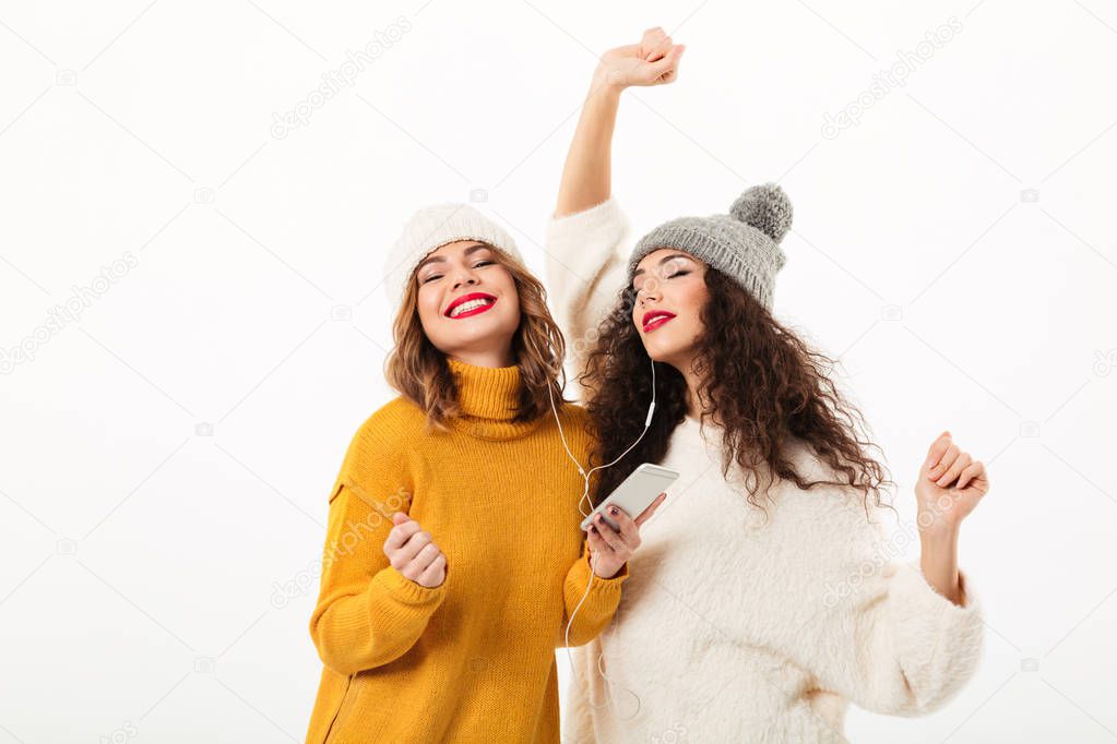 Two pleased girls in sweaters and hats dancing together