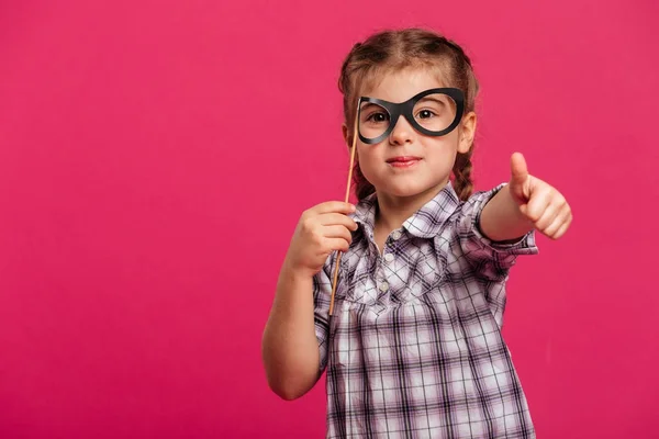 Ragazzina allegra bambino tenendo occhiali falsi mostrando pollici in su . — Foto Stock