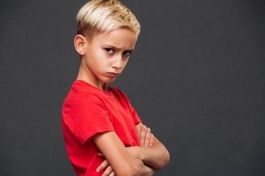 Photo of angry little boy child standing isolated over grey background. Looking camera with arms crossed.