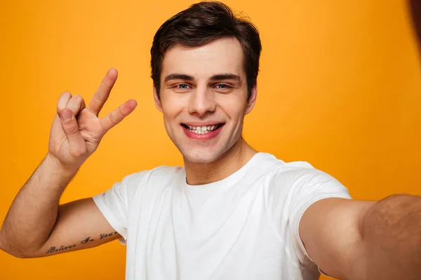 Homem sorridente em t-shirt fazendo selfie enquanto mostra gesto de paz — Fotografia de Stock