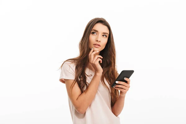 Mujer morena pensativa en camiseta sosteniendo smartphone y mirando hacia otro lado — Foto de Stock