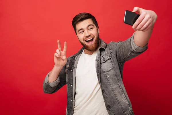 Hombre barbudo alegre haciendo selfie en el teléfono inteligente y mostrando paz — Foto de Stock