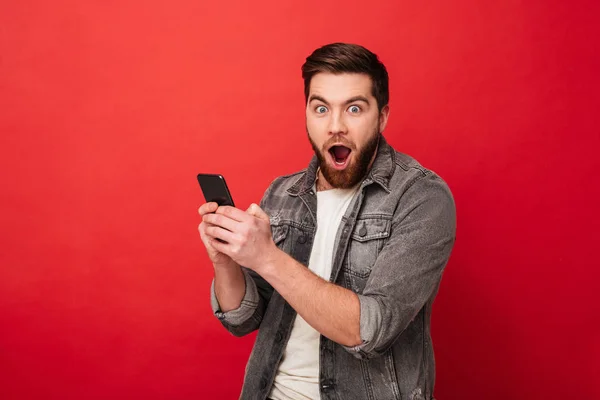 Здивований щасливий бородатий чоловік тримає смартфон і дивиться на камеру — стокове фото