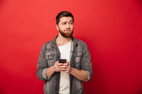 Confused görüntüsünü smartphone tutarak ve uzağa bakarak adam sakallı — Stok fotoğraf