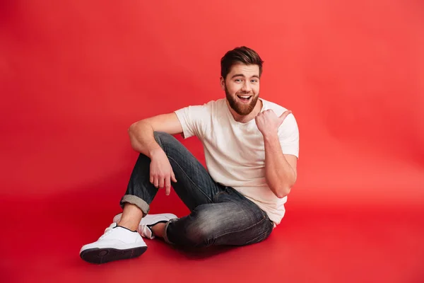 Smiling bearded man sitting on floor pointing. — Stock Photo, Image