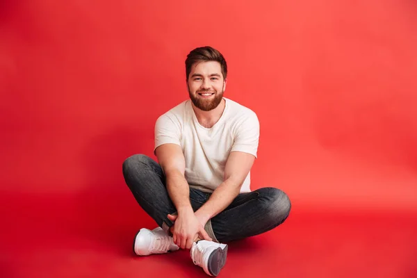Бородатий чоловік сидить на підлозі ізольовано на червоному тлі — стокове фото