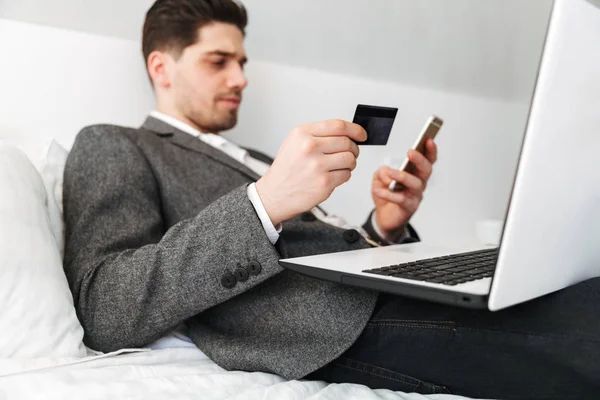 Foto des smarten Mannes im Anzug, der im Bett liegt, während er online Witze bezahlt — Stockfoto