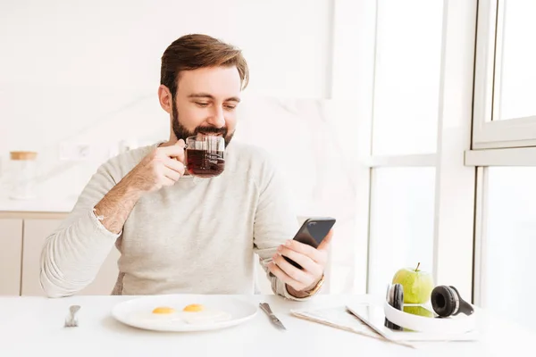 Bearded man in casual shirt drinking tea and using smart phone, — Stock Photo, Image