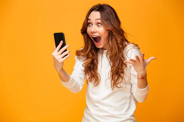 Mulher morena chocada em suéter segurando smartphone e se alegra — Fotografia de Stock