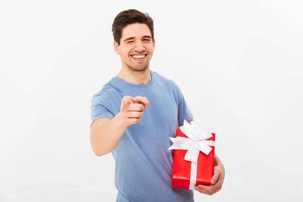 Smilende mand i t-shirt med gave, mens han peger på kameraet - Stock-foto