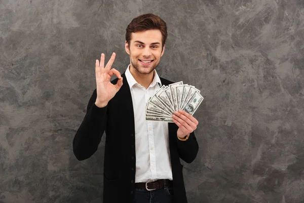 Šťastný mladý podnikatel drží peníze při pohledu kamery zobrazeno pořádku gesto. — Stock fotografie