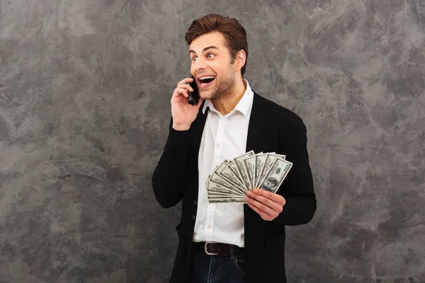 Jovem empresário alegre segurando dinheiro olhando para o lado falando por telefone celular . — Fotografia de Stock