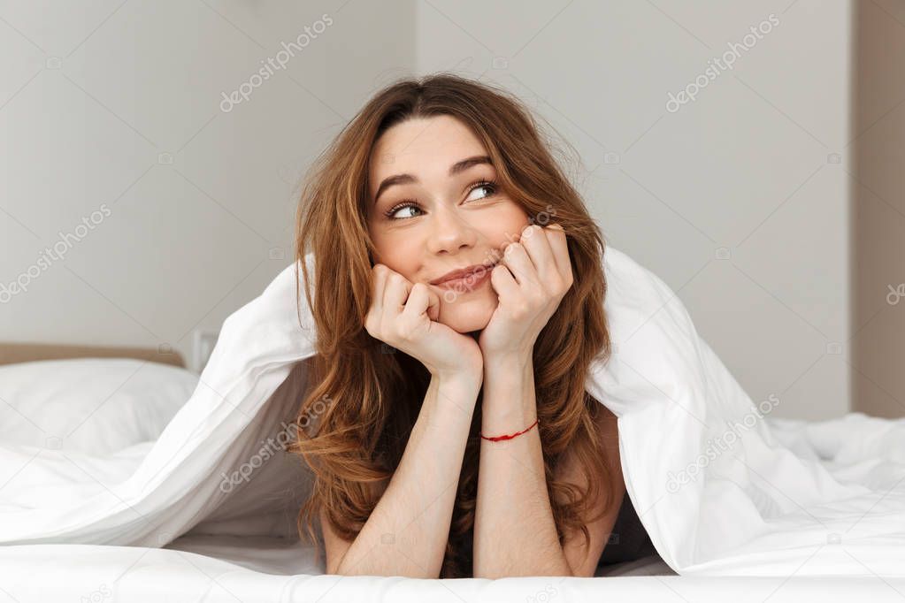 Portrait of alluring young woman lying in bed under blanket at h