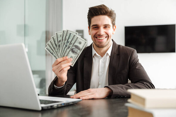 Cheerful young businessman showing money using laptop computer.