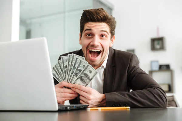 Ενθουσιασμένος νέος επιχειρηματίας δείχνει χρήματα που χρησιμοποιούν φορητό υπολογιστή. — Φωτογραφία Αρχείου