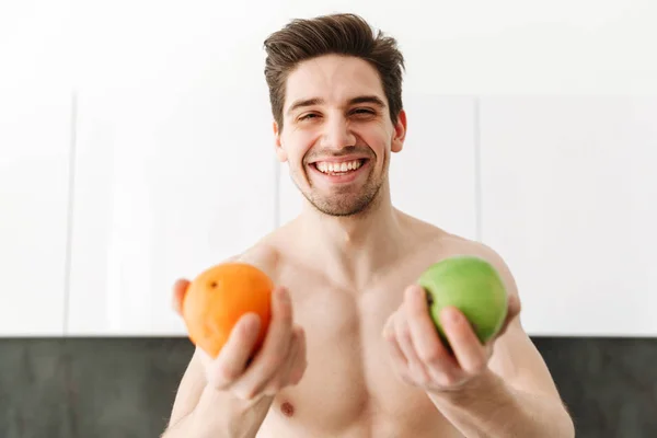 Happy cheerful naked man sportsman showing orange and apple — Stock Photo, Image