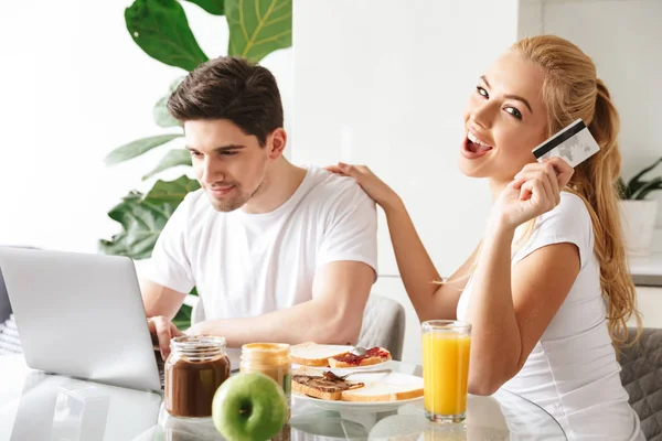 Sorrindo jovem casal amoroso tem um café da manhã usando computador portátil segurando cartão de crédito . — Fotografia de Stock