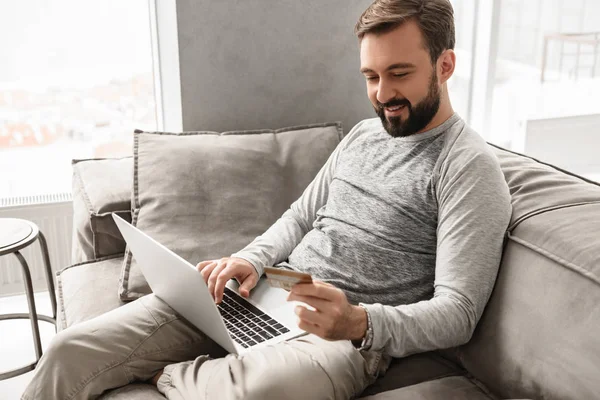 Piacevole uomo moderno 30s in abbigliamento casual seduto sul divano in soggiorno — Foto Stock