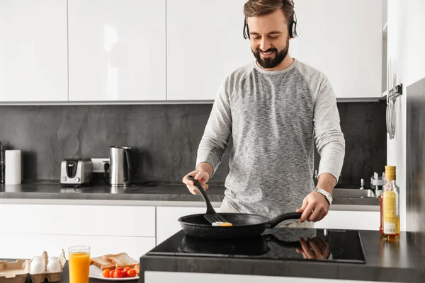 Cheerful man 30s wearing casual clothing frying eggs for breakfa — Stock Photo, Image