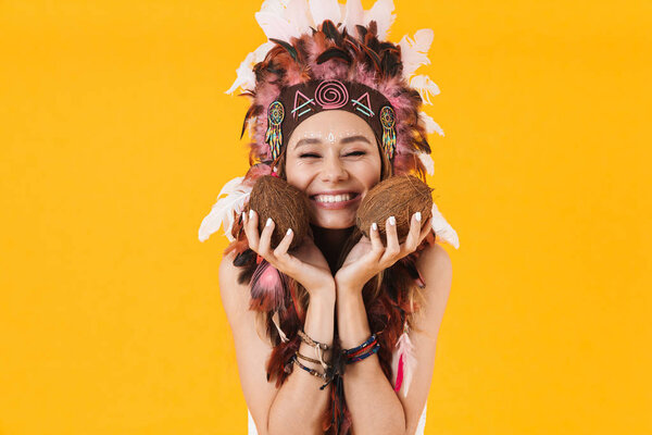 Portrait of attractive smiling woman in headdress of feathers holding coconuts with eyes closed isolated over yellow background
