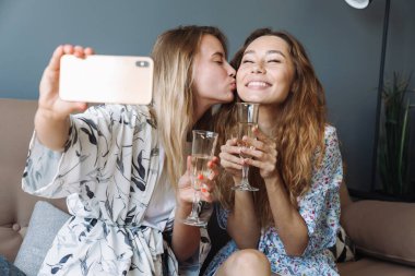 Evde şampanya içen iyimser kadınlar cep telefonlarıyla selfie çekiyorlar..