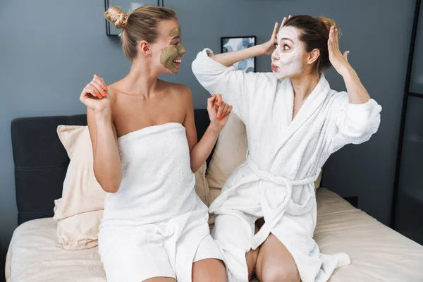 Vidám fiatal nők vigyáznak a bőrükre arcmaszkkal ülnek az ágyon.. — Stock Fotó