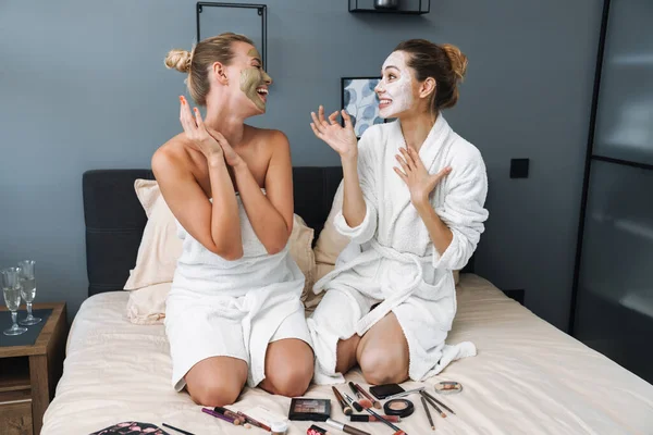 Allegro giovani donne prendersi cura della loro pelle con maschera siedono sul letto . — Foto Stock
