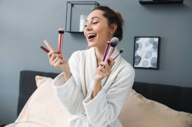 Neşeli, iyimser, mutlu, elinde makyaj fırçası tutan genç bir kadın..