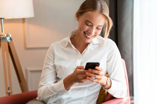 Χαρούμενη όμορφη ξανθιά επιχειρηματίας κάθεται σε εσωτερικούς χώρους στο καφέ χρησιμοποιώντας το κινητό τηλέφωνο. — Φωτογραφία Αρχείου