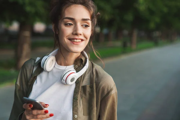 Imagen de mujer alegre sosteniendo teléfono inteligente y caminando en verde pa — Foto de Stock