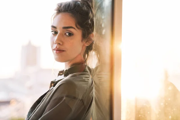 Imagen de la encantadora mujer morena de pie sobre la puerta de cristal mientras —  Fotos de Stock