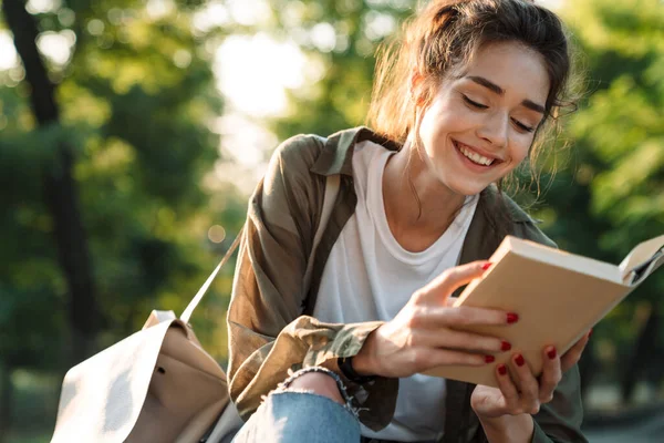 Imagen de mujer atractiva sonriendo y leyendo libro en parque verde — Foto de Stock