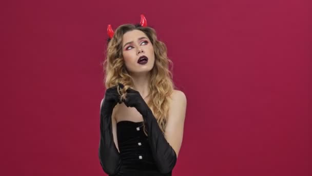 カーニバルの衣装を着た美しい悪魔の女性は何かを考え 赤い壁の背景に隔離された彼女の髪に触れながらアイデアジェスチャーを示します — ストック動画