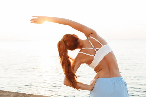 Piękna młoda ruda sport kobieta zrobić rozciągające się ćwiczenia na zewnątrz na plaży rano. — Zdjęcie stockowe