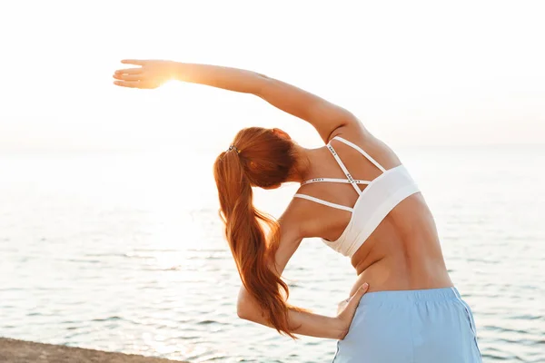 Schöne junge rothaarige Sportlerin macht morgens Dehnübungen im Freien am Strand. — Stockfoto