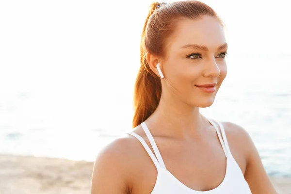 Piacevole bella giovane rossa donna sportiva seduta all'aperto in spiaggia al mattino ascoltando musica con gli auricolari . — Foto Stock