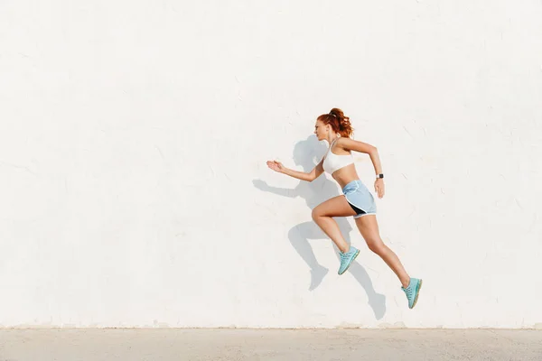 Imagen de la joven pelirroja corriendo a lo largo de la pared blanca mientras hace — Foto de Stock