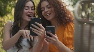 Çekici, neşeli genç kızlar. Parkta otururken birbirlerine gülümseyen ve akıllı telefonlarıyla bir şeyler gösteren arkadaşlar.
