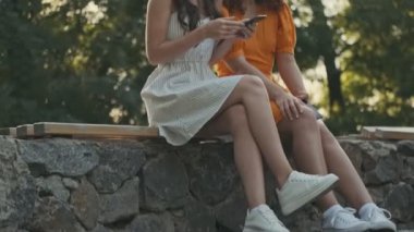 Attractive cheerful young girl laughing loudly while using smartphone and her friend smiling but trying not to laugh while sitting in the park
