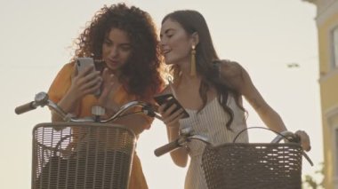 Beautiful young girls friends speaking and watching something on their smartphones while sitting on their bicycles outdoors at the street