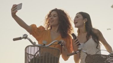 Neşeli, genç kız arkadaşlar, sokakta bisikletleriyle otururken akıllı telefonlarında gülümseyip selfie çekiyorlar.