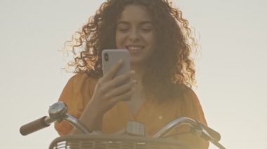 Neşeli, genç, kıvırcık saçlı, gülümseyen ve akıllı telefon kullanan bir kadın sokakta bisikletiyle oturuyordu.