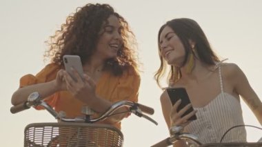 Neşeli, güzel genç kızlar gülümseyen ve akıllı telefonlarını kullanan arkadaşları sokakta bisikletlerinin üzerinde oturuyorlar.