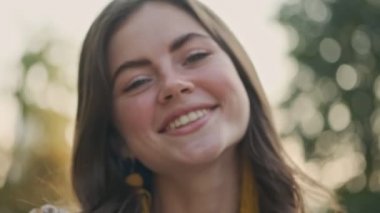 Parkta dikilirken gülümseyen ve saçlarıyla oynayan neşeli, genç esmer kadının yakın görüntüsü.