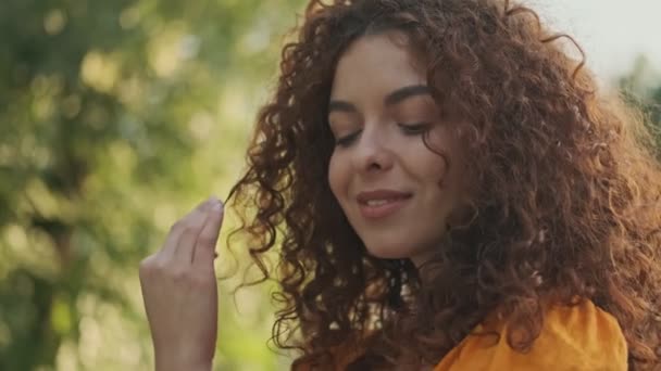 在公园休息的时候 穿着橙色衣服的漂亮可爱的红头发女人笑着玩着卷曲的头发 — 图库视频影像