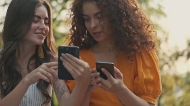 Cute cheerful young girls friends showing to each other something on their smartphones and laughing while sitting in the park